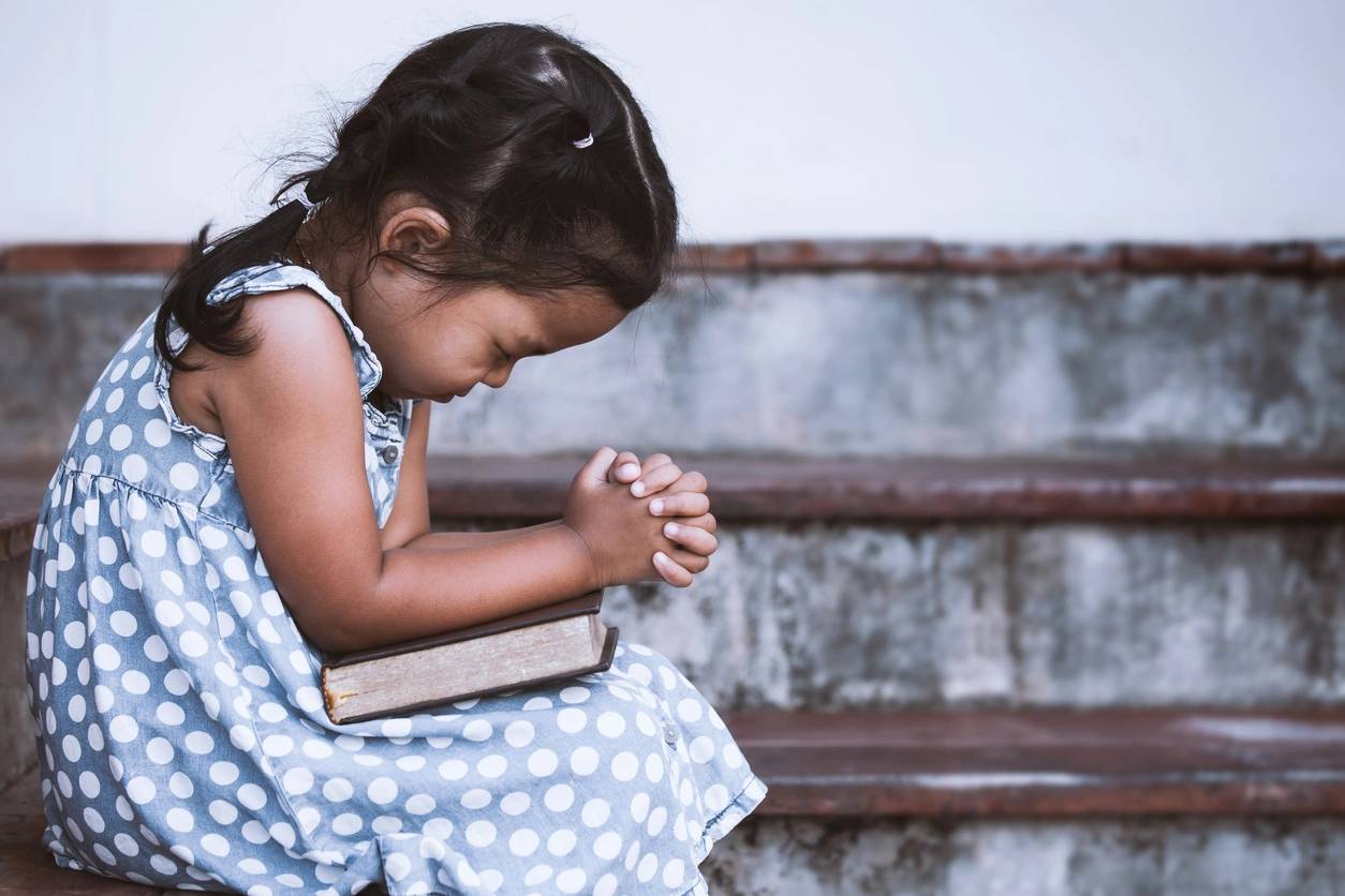 A young girl kneeling in prayer with folded hands and a holy book resting in her lap, in a serene and peaceful setting.