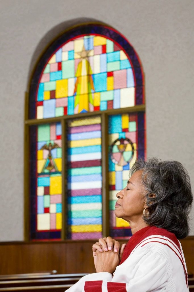 A woman praying in a church, hands clasped and eyes slightly looking upward, with a stained glass window behind her, symbolizing how to pray